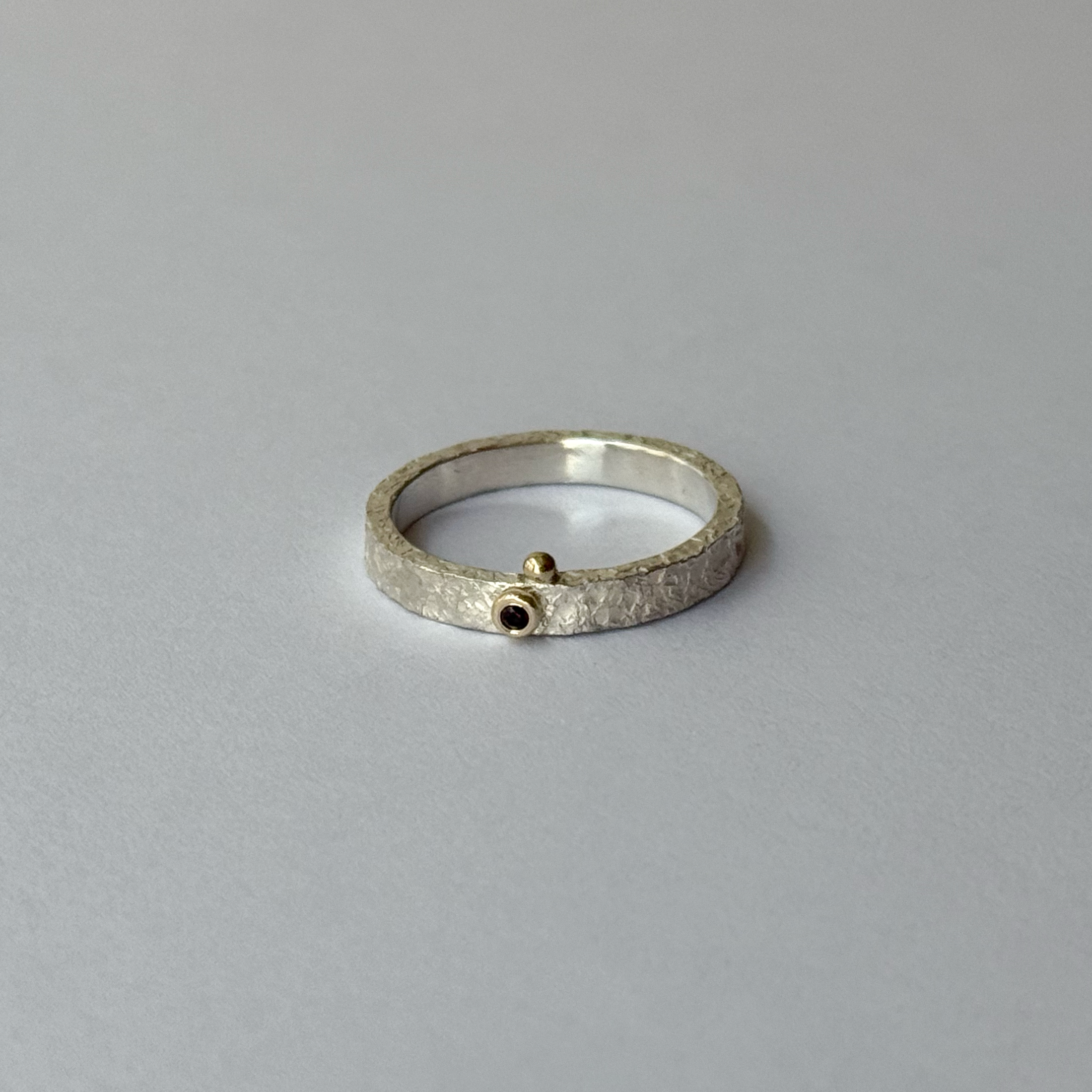 A silver ring with a red garnet in the center, decorated with gold bubbles, placed on a light background.