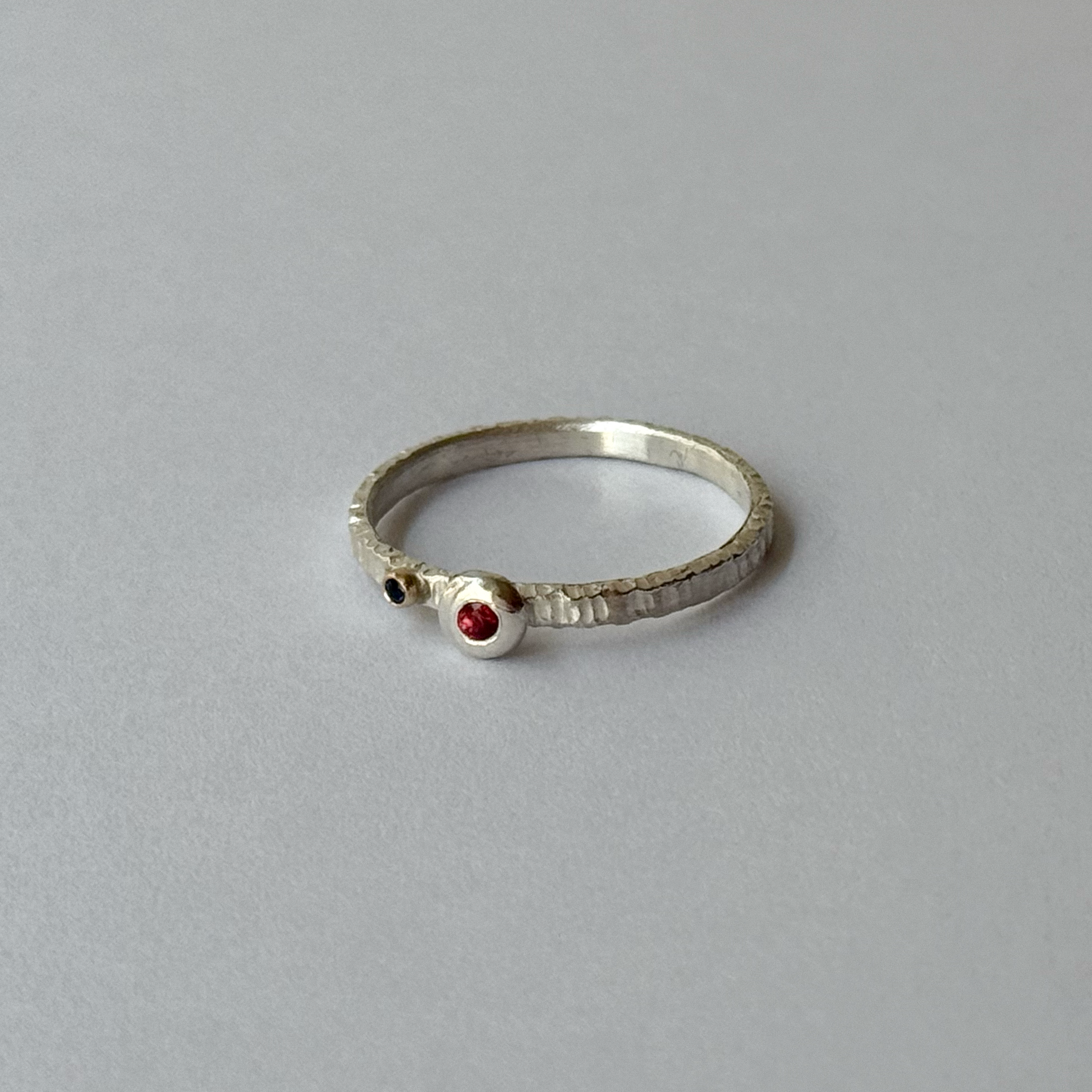 A silver ring with orange and blue sapphires set on the top, placed on a grey background.