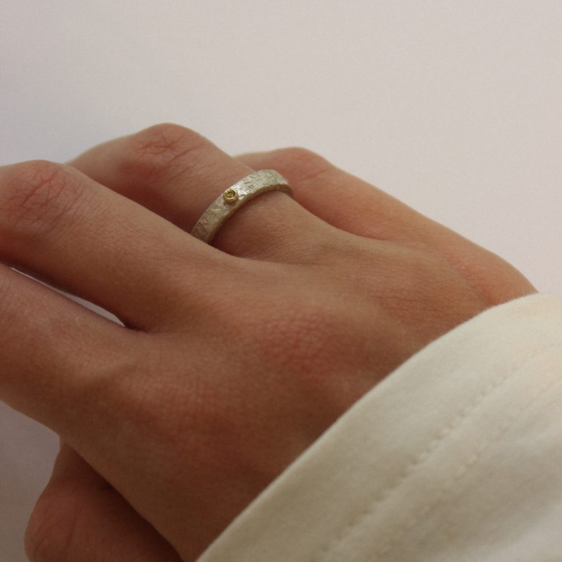 A close-up image of a hand holding a narrow silver and yellow diamond ring with a gold sphere on top.