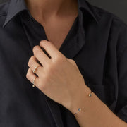 A close-up of a person wearing delicate gold rings and a thin gold bracelet with small dark blue stones, paired with a black button-down shirt.