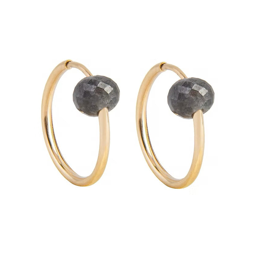 A pair of gold hoop earrings with a small, faceted dark gray diamond bead attached near the top right of each hoop, shown against a plain white background.