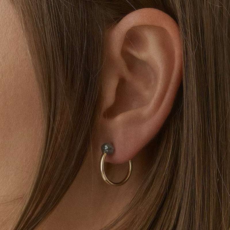 A close-up of a person's ear wearing a small gold hoop earring with a tiny dark gray faceted diamond on the lobe.
