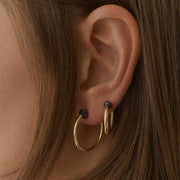 A close-up of a person's ear wearing two gold hoop earrings, one larger hoop and one smaller stacked hoop, with small dark faceted diamonds on the studs.