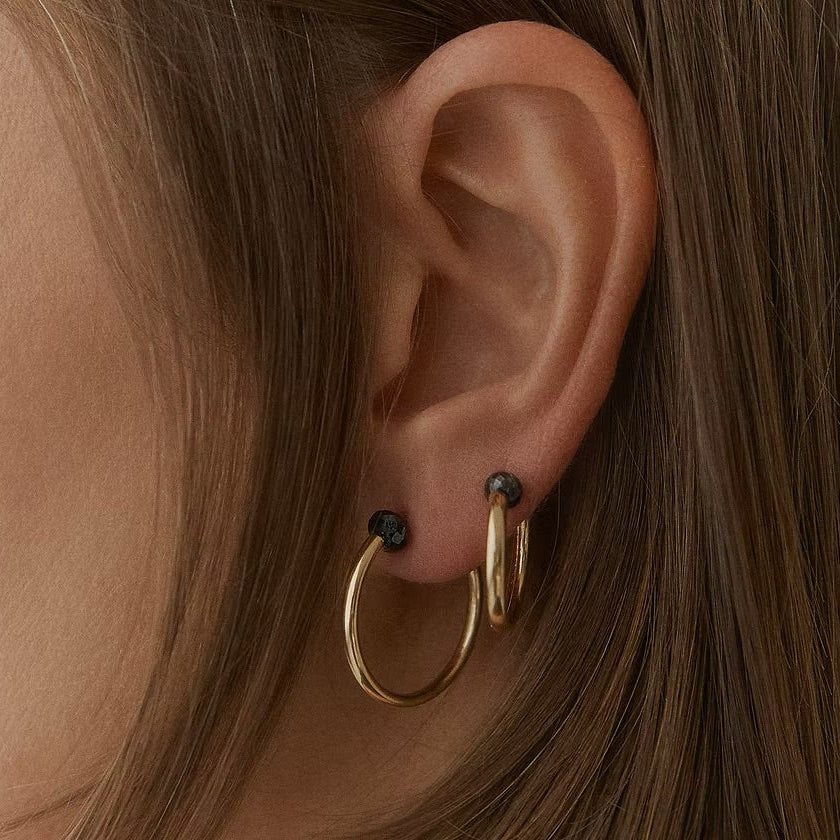 A close-up of a person's ear wearing two gold hoop earrings, one larger hoop and one smaller stacked hoop, with small dark faceted diamonds on the studs.