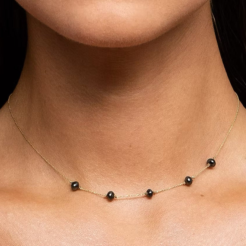 A close-up of a model's neck and collarbone wearing a delicate gold chain necklace with five small, evenly spaced black faceted diamond beads.