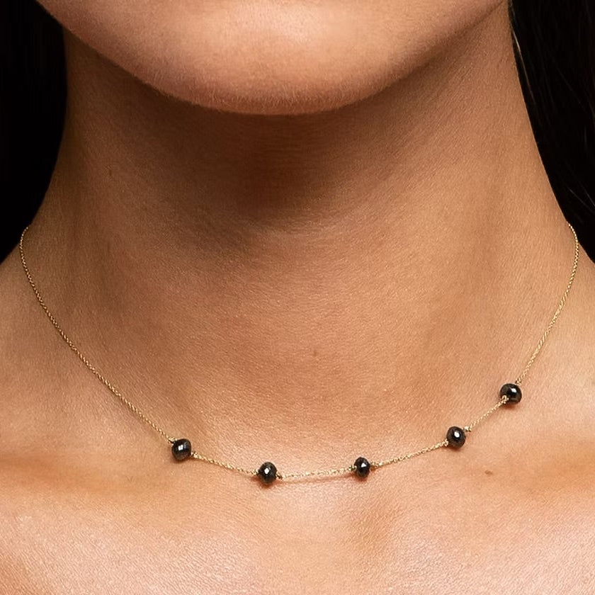 A close-up of a model's neck and collarbone wearing a delicate gold chain necklace with five small, evenly spaced black faceted diamond beads.