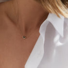 A close-up of a model's neck and chest wearing a delicate gold chain necklace with a small, dark oval pendant, styled with a white collared shirt.