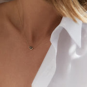 A close-up of a model's neck and chest wearing a delicate gold chain necklace with a small, dark oval pendant, styled with a white collared shirt.