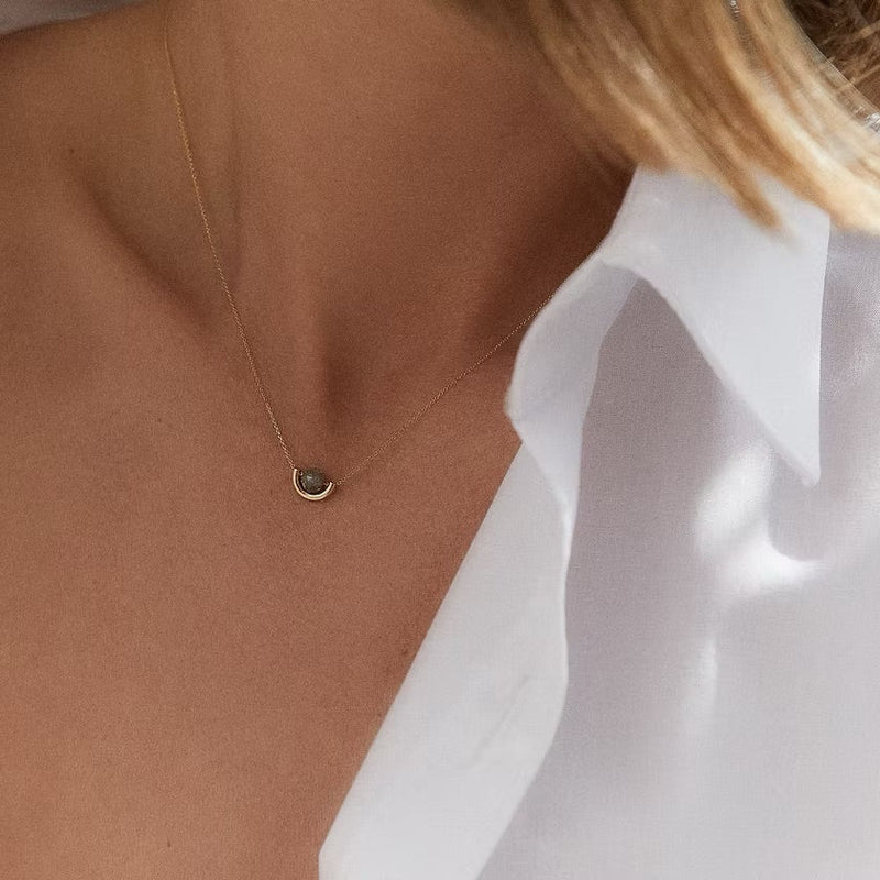 A close-up of a model's neck and chest wearing a delicate gold chain necklace with a small, dark oval pendant, styled with a white collared shirt.