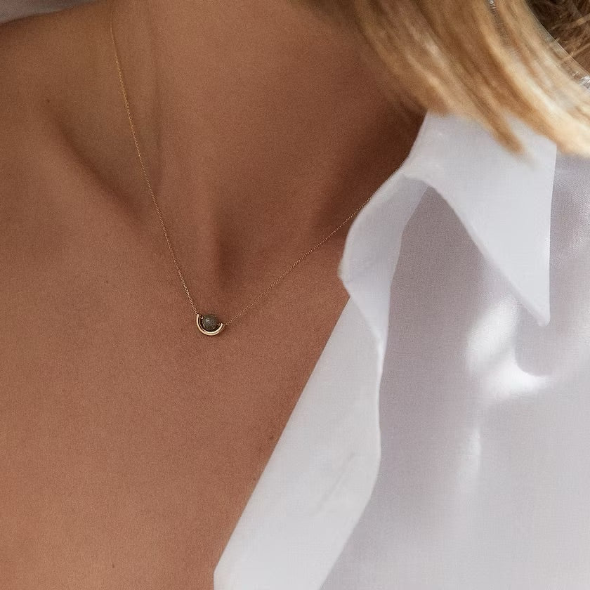 A close-up of a model's neck and chest wearing a delicate gold chain necklace with a small, dark oval pendant, styled with a white collared shirt.