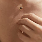 Close-up of a person's chest and hand wearing a delicate gold necklace with a round dark gemstone pendant.