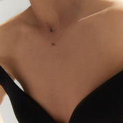 A close-up of a model's neck and chest wearing a delicate layered necklace with two small dark faceted diamond pendants on fine chains, paired with a black strapless top.