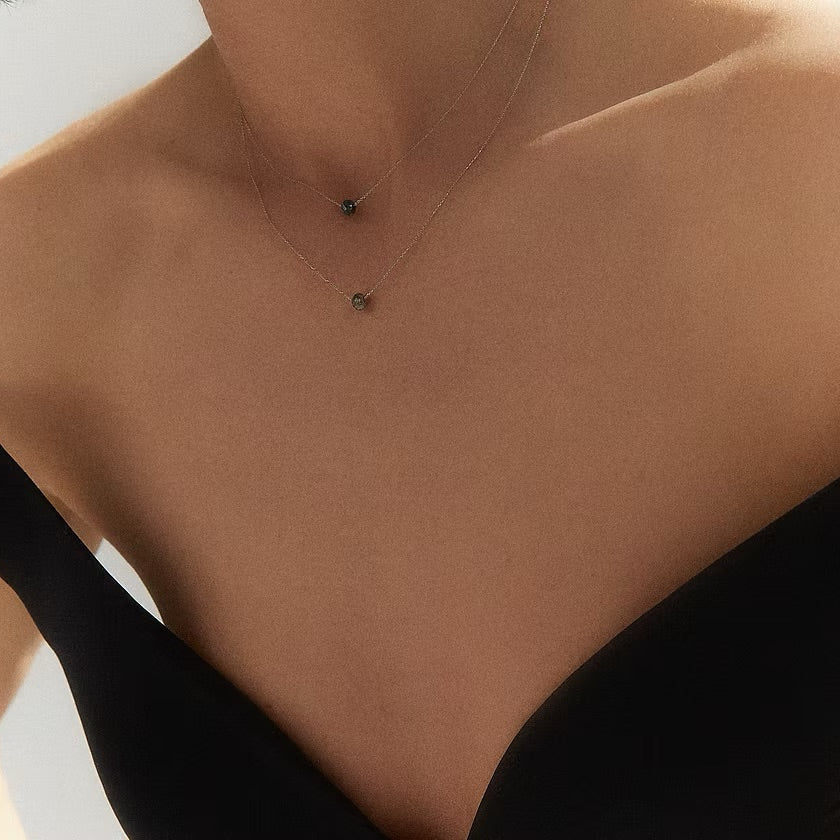 A close-up of a model's neck and chest wearing a delicate layered necklace with two small dark faceted diamond pendants on fine chains, paired with a black strapless top.