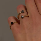 A close-up of a hand wearing two slim open rings with dark faceted diamonds, against a plain gray background.