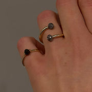 A close-up of a hand wearing two slim open rings with dark faceted diamonds, against a plain gray background.