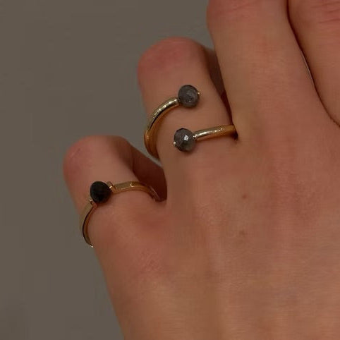 A close-up of a hand wearing two slim open rings with dark faceted diamonds, against a plain gray background.