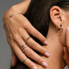 A close-up lifestyle shot of a model wearing multiple delicate gold rings on her fingers and ear, including a small hoop earring with a black diamond. 
