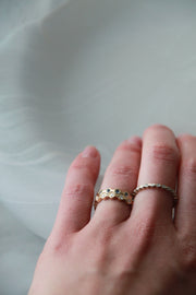 A close-up of a hand wearing two delicate rings: a gold band with circular cutout detailing and a thin silver band with small clear stones, set against a soft, light gray background.
