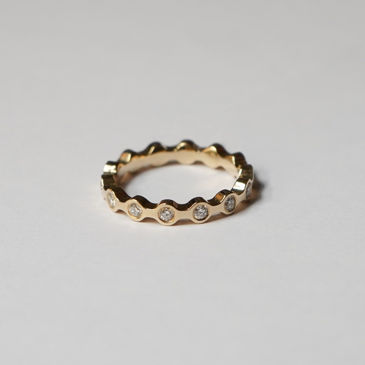 A gold eternity-style ring with a wavy, scalloped edge and small clear gemstones set along the front, photographed on a clean light gray background.