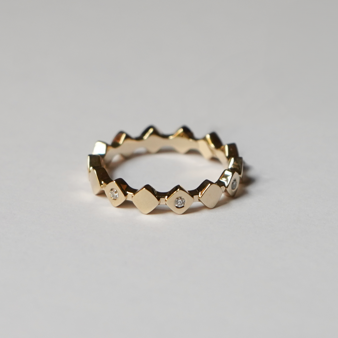A gold geometric ring with alternating diamond and triangular links, accented by small diamonds, photographed on a neutral light gray background.