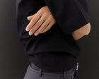 A close-up of a person's hand wearing three delicate gold rings, styled with a black blazer and gray trousers against a dark background.