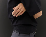 A close-up of a person's hand wearing three delicate gold rings, styled with a black blazer and gray trousers against a dark background.