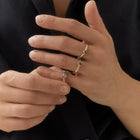 A close-up of a person's hands wearing three delicate gold rings with small grey diamonds, set against a dark navy shirt.
