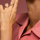 Close-up of a person wearing two delicate yellow gold and grey diamonds rings on their fingers. The person is wearing a pink leather collared shirt against a solid dusty rose background.