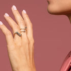 Close-up of a person wearing two delicate yellow gold and grey diamonds rings on their fingers. The person is wearing a pink leather collared shirt against a solid dusty rose background.