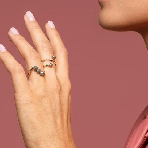 Close-up of a person wearing two delicate yellow gold and grey diamonds rings on their fingers. The person is wearing a pink leather collared shirt against a solid dusty rose background.