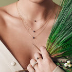 A close-up lifestyle shot of a model wearing delicate layered gold necklaces with small dark bead pendants and rings, holding a green palm frond against a neutral background.