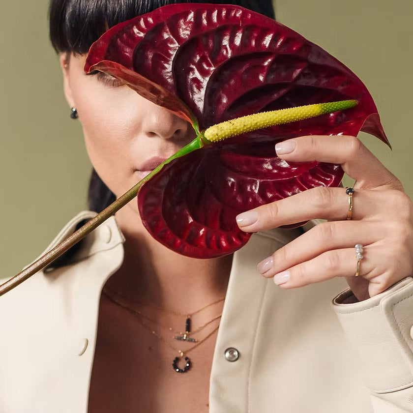 Person holding a large red flower against a plain background. Her hands is adorned with gold & rax diamonds rings.