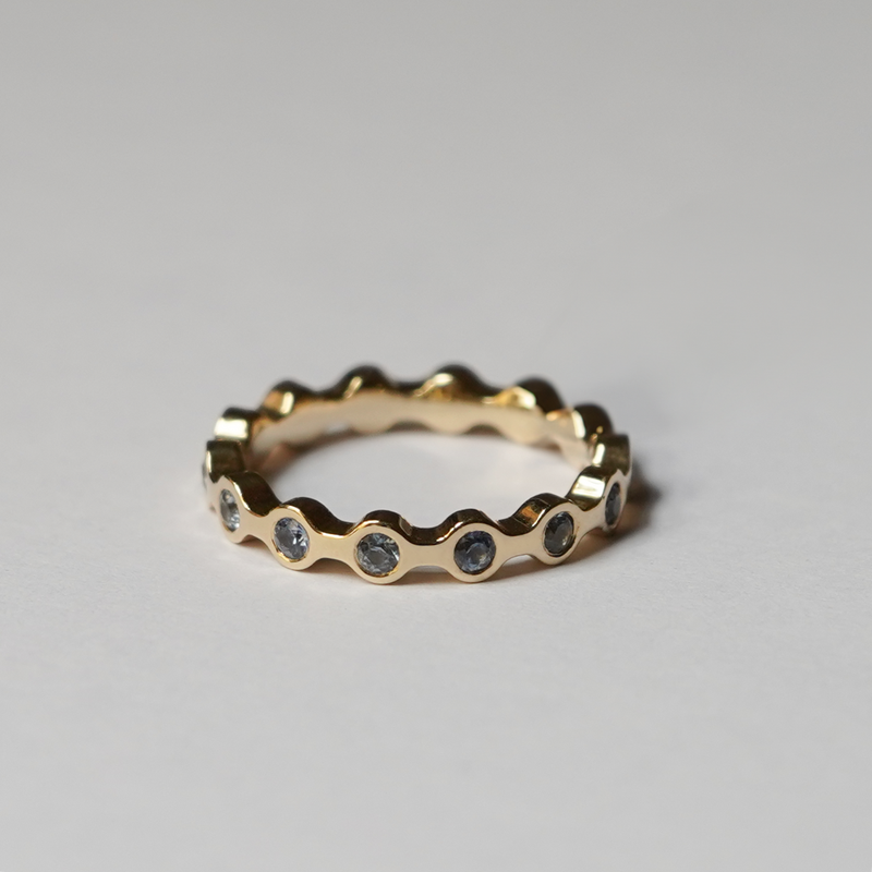 A gold eternity ring with a wavy, scalloped edge and small round blue sapphires set along the band, photographed on a neutral light gray background.