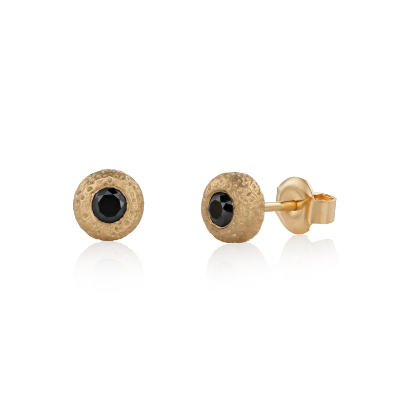 A pair of gold round stud earrings with a textured finish and a central black round diamond, shown on a white background with soft reflections.