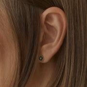 A close-up of a person's ear wearing a small dark stud earring with a yellow gold bezel setting and a tiny clear center stone, surrounded by brown hair.
