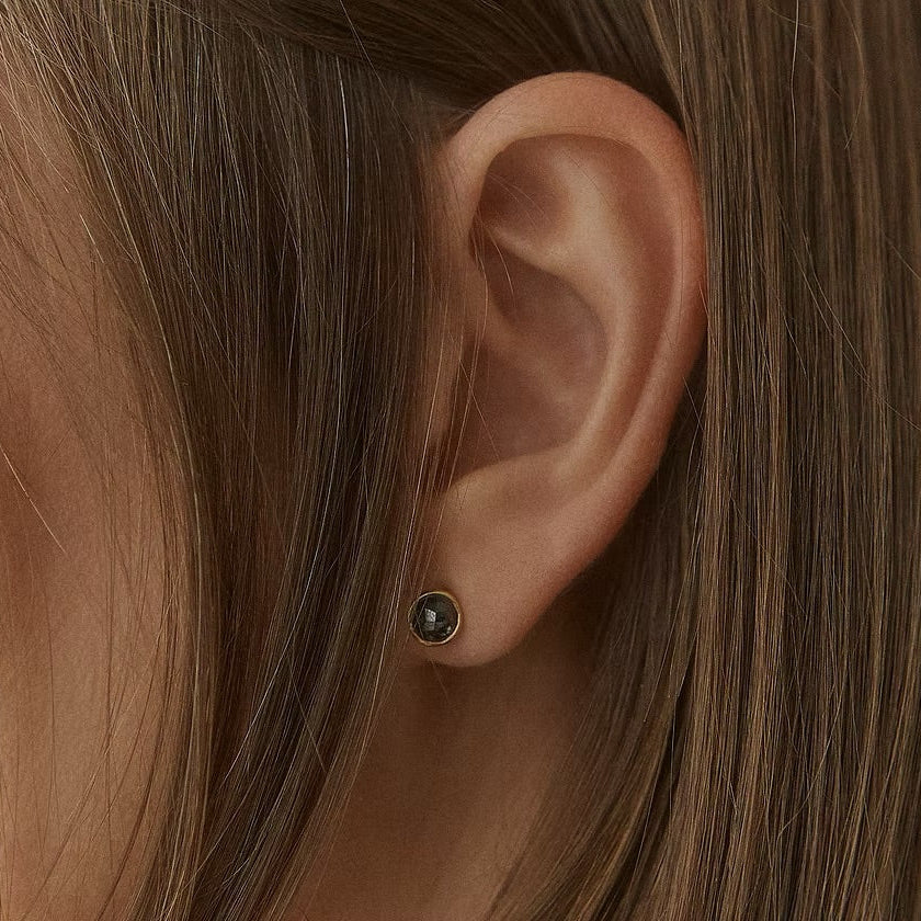 A close-up of a person's ear wearing a small dark stud earring with a yellow gold bezel setting and a tiny clear center stone, surrounded by brown hair.