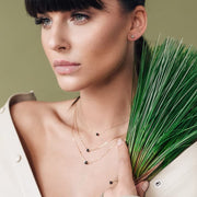A close-up lifestyle shot of a model wearing delicate gold layered necklaces with small black beads, holding a green grass-like plant against a solid olive green background.