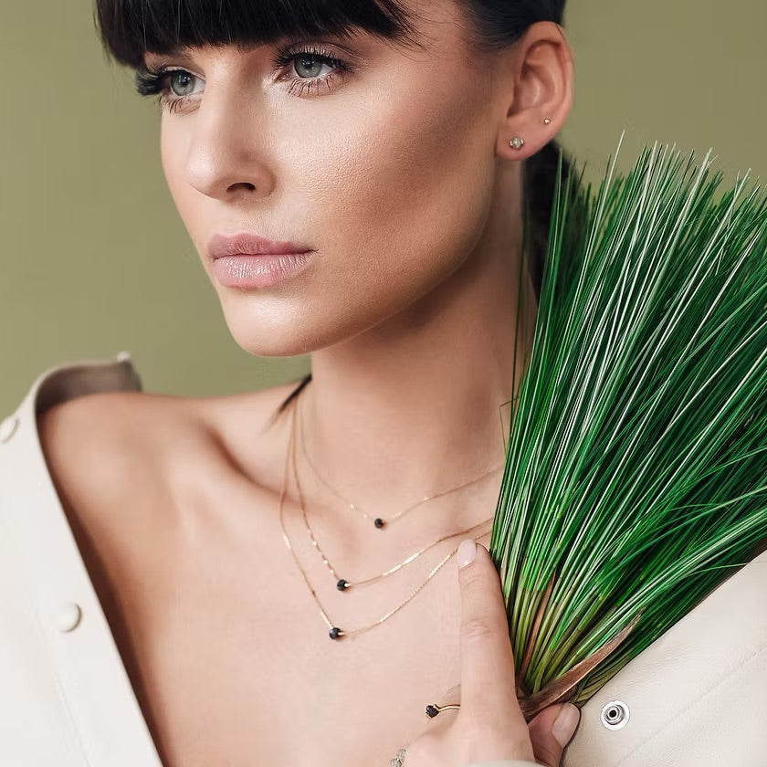 A close-up lifestyle shot of a model wearing delicate gold layered necklaces with small black beads, holding a green grass-like plant against a solid olive green background.