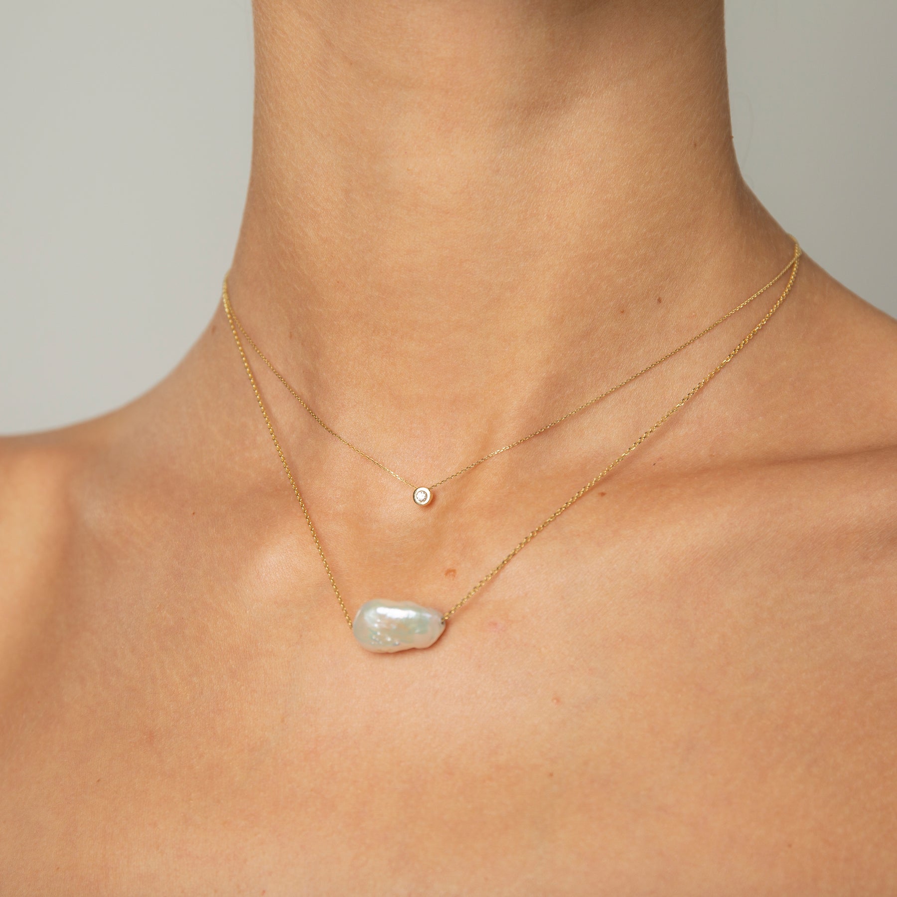 Woman wearing two minimal design yellow gold necklaces. One is with diamond, the other is with baroque pearl.