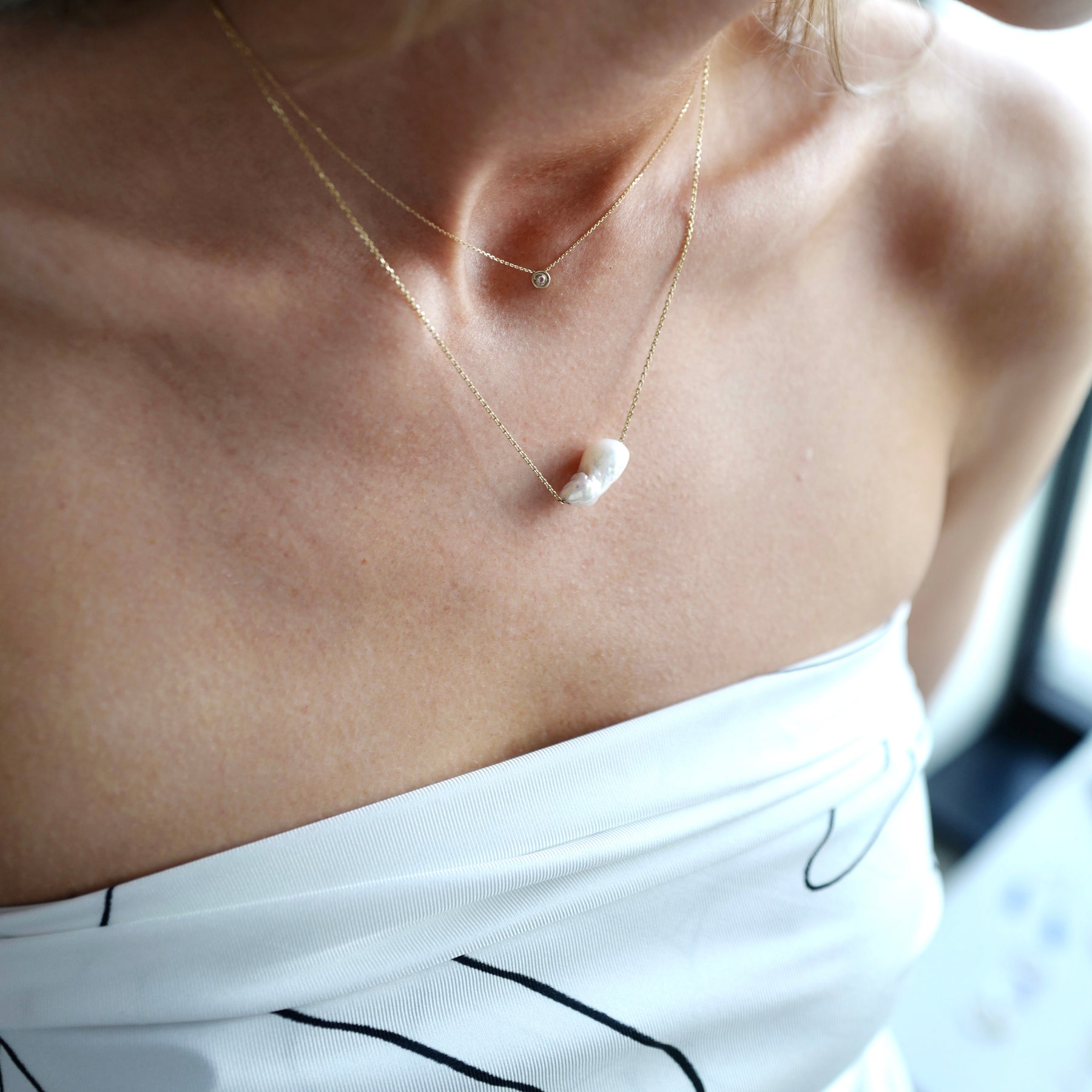Woman wearing two minimal design yellow gold necklaces. One is with diamond, the other is with baroque pearl.