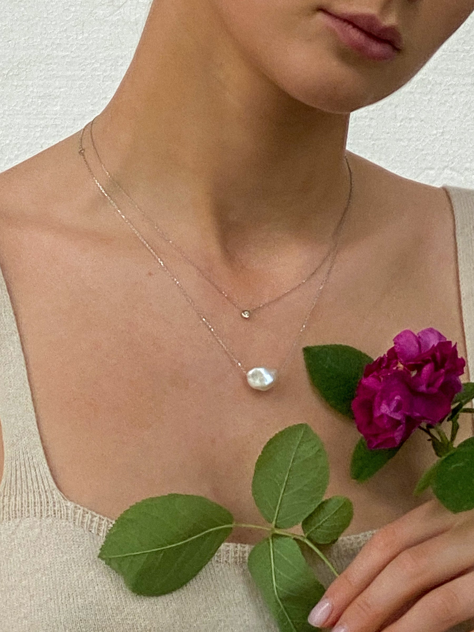 Woman wearing two minimal design yellow gold necklaces. One is with diamond, the other is with baroque pearl.