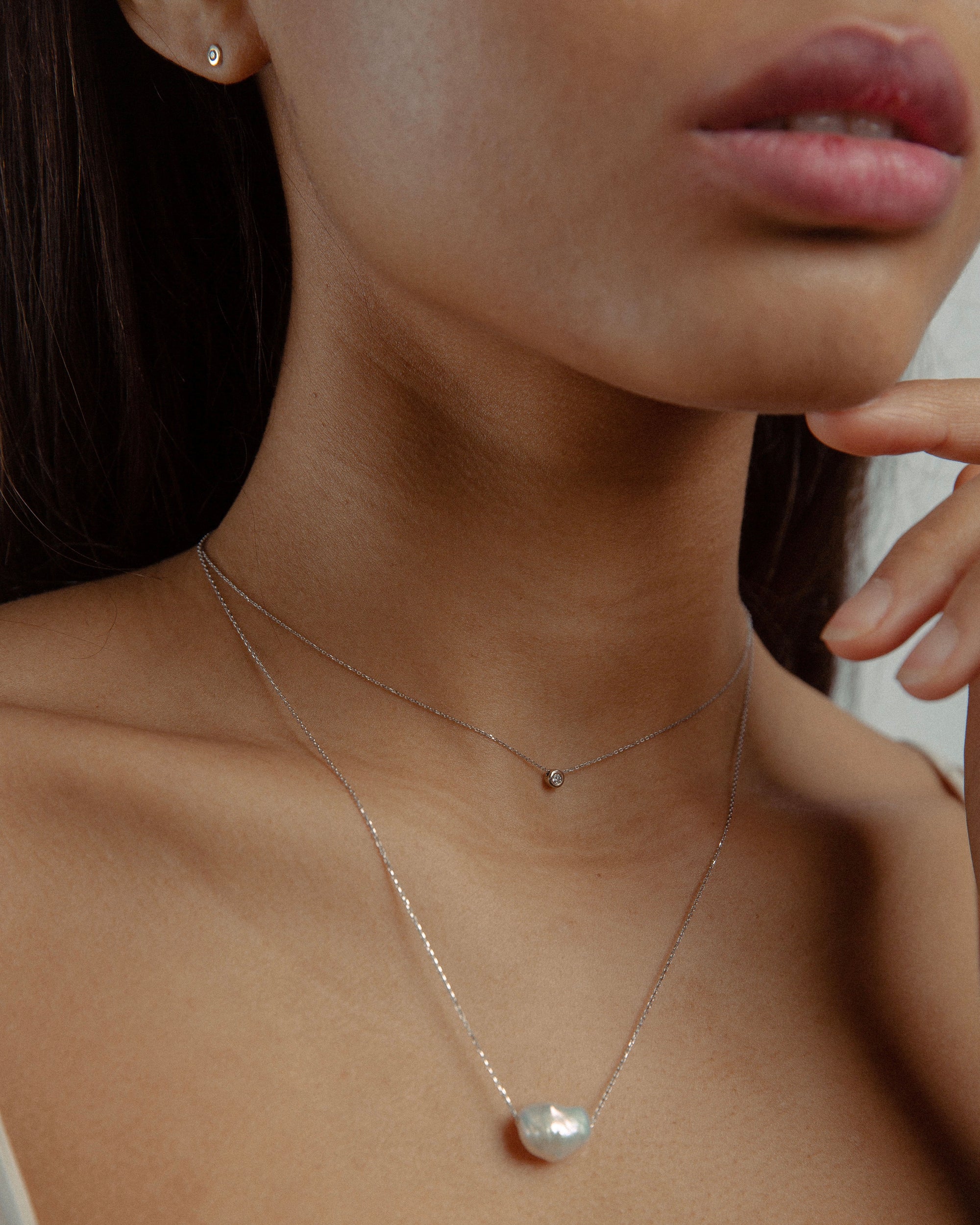 Woman wearing two minimal design yellow gold necklaces. One is with diamond, the other is with baroque pearl.